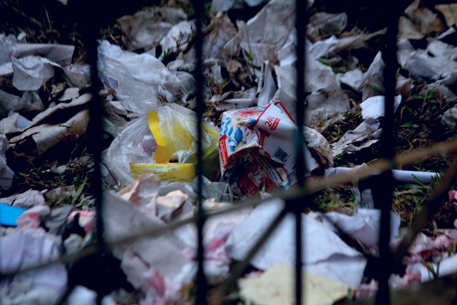 A close-up view through black metal garden fencing showing a pile of discarded garden waste and rubbish, including crumpled paper, plastic bags, and packaging materials on the ground. The background r