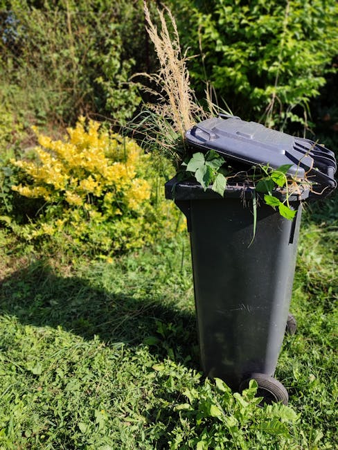 A close-up view through black metal garden fencing showing a pile of discarded garden waste and rubbish, including crumpled paper, plastic bags, and packaging materials on the ground. The background r