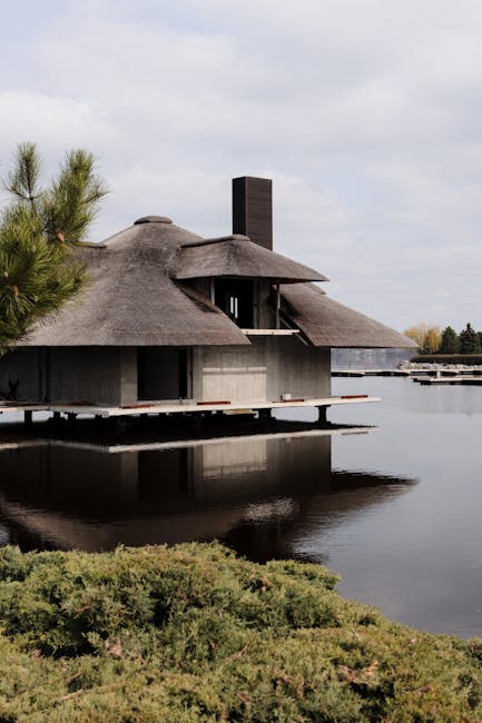 A modern, eco-friendly house with a thatched roof situated on the edge of a calm body of water, offering a scenic view of the surrounding landscape. The house features dark, vertical chimney structure